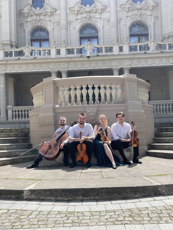 Kobieta i 3 mężczyzn - członkowie zespołu Vivenda String Quartet z instrumentami smyczkowymi siedzący przed budynkiem